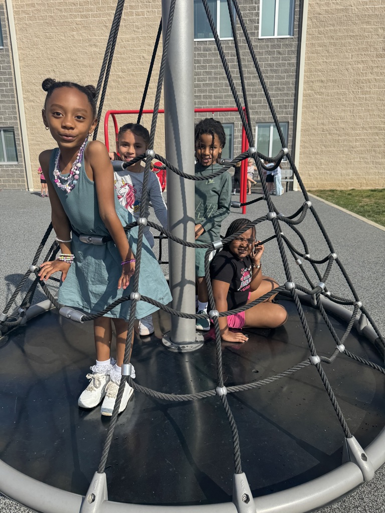 Four young girls play on a circular, netted playground structure in front of a building.