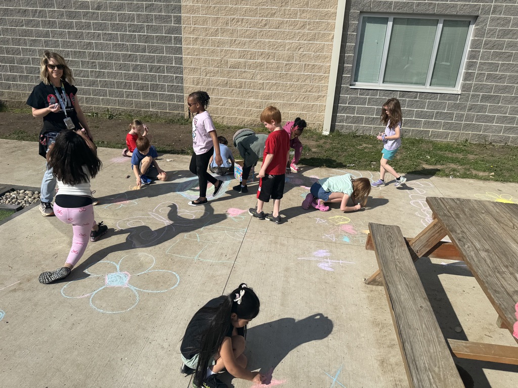 Kids are outdoors drawing pictures in chalk on the ground near a building.