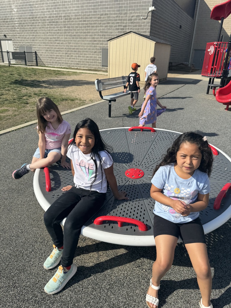 Three children sit on a round ride in a playground. One is sitting on the edge, while the others are seated. Other children are playing in the background.