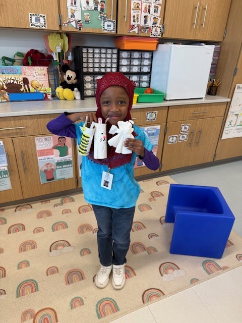 A young child stands in a classroom, holding white objects. They wear a blue shirt and red head covering. Behind them, wooden cabinets and a refrigerator are visible.