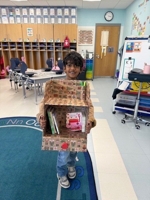A child stands in a classroom, holding a box filled with books and other items. The room features chairs, tables, and a clock on the wall.