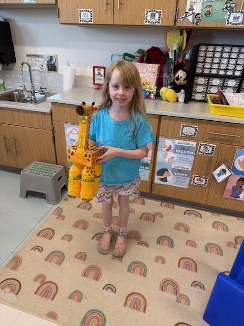 Young girl in a blue shirt holding a giraffe-shaped object in a classroom with wooden cabinets and a sink.