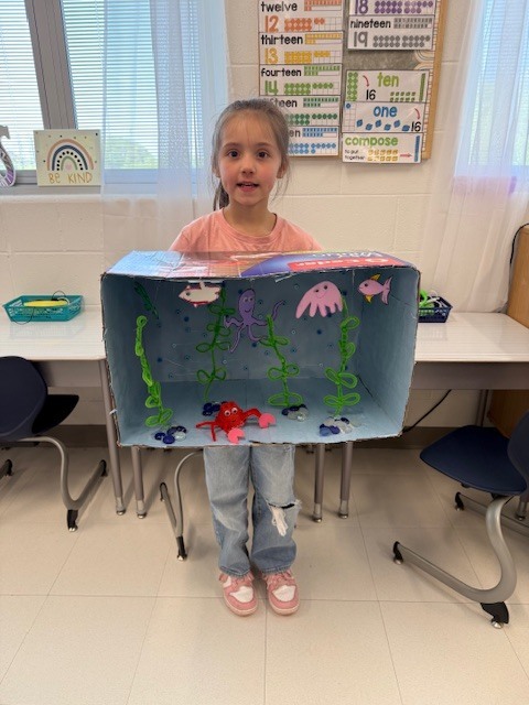 A child stands in a classroom, holding a box aquarium with fish and plants inside.