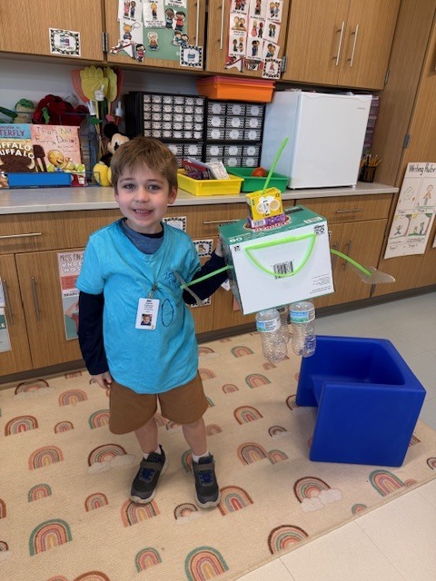 A child in a blue shirt and shorts stands in a classroom, holding a cardboard robot in one hand.