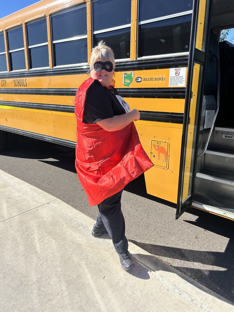 A woman in a red cape and black shirt stands next to a yellow school bus.