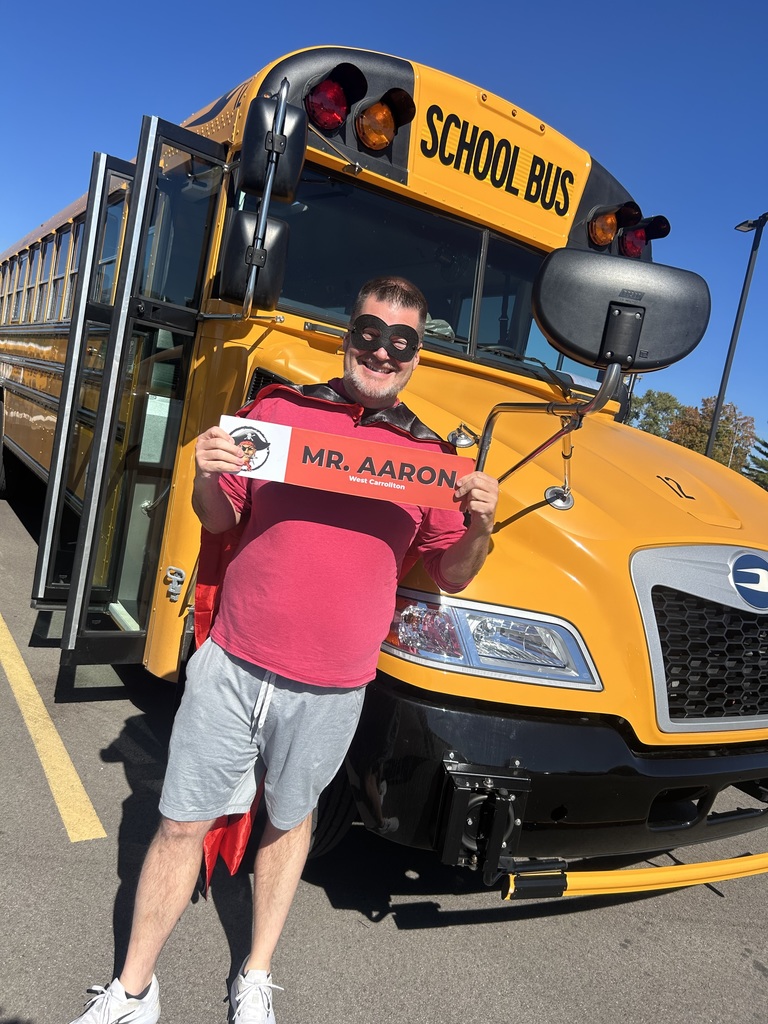 A person wearing a red cape and mask holds a card with "Mr. Aaron" on it, standing before a yellow school bus.