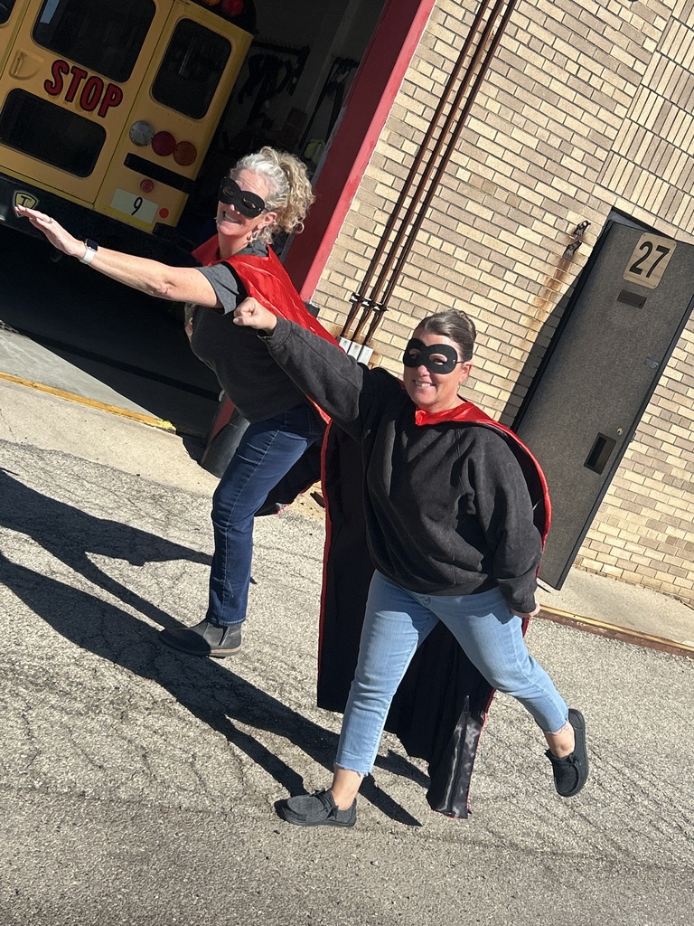 Two people wear superhero capes and sunglasses in front of a yellow school bus and a brick building.
