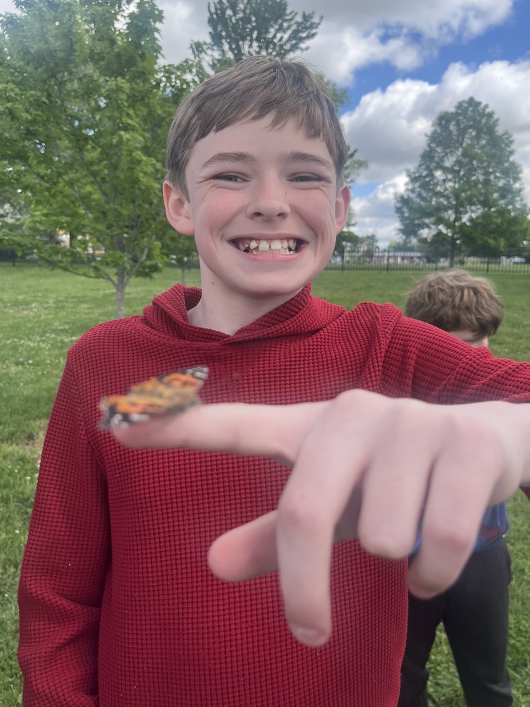 A boy has a butterfly resting on his finger. Trees and grass in the background.