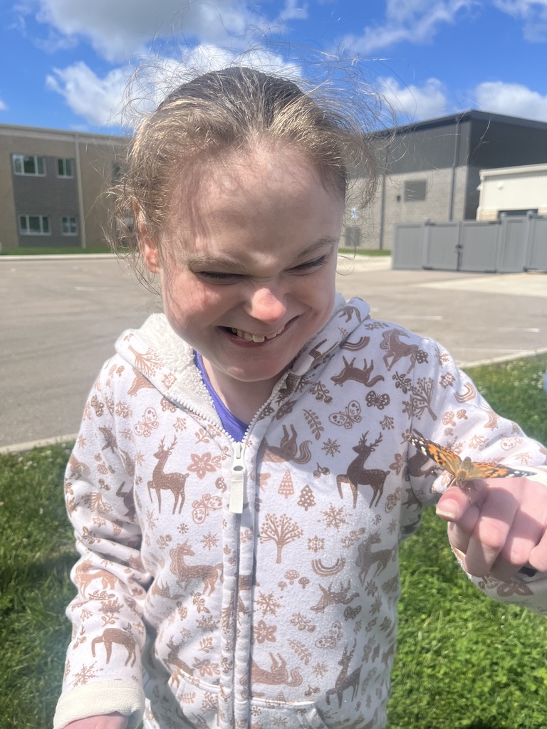 A young girl smiles, holding a butterfly. Behind her is a grassy area and a building.