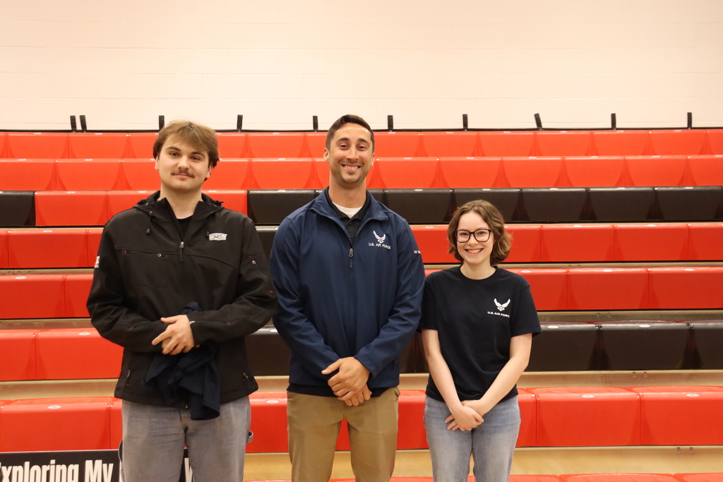 Three individuals in black, blue, and white shirts stand in a gym with red seating.