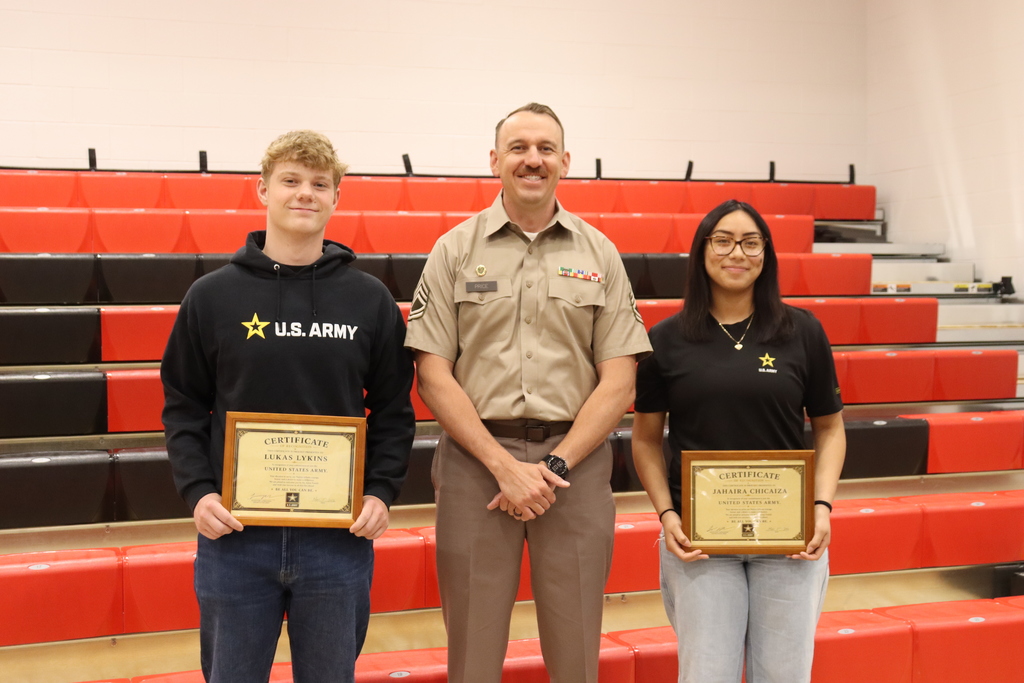 Three people stand in a gym. The man in the center wears a beige uniform, while the two others wear black shirts.