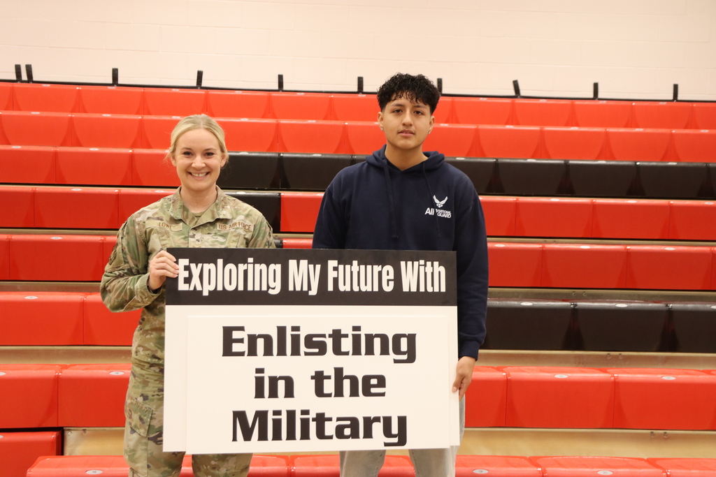 Two individuals stand in a gymnasium with a red bleacher background, holding a sign with the text "Exploring My Future With Enlisting in the Military."
