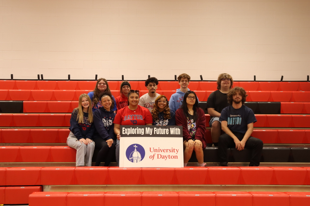 Group of people sit in a red stadium seat. One holds a sign for University of Dayton.