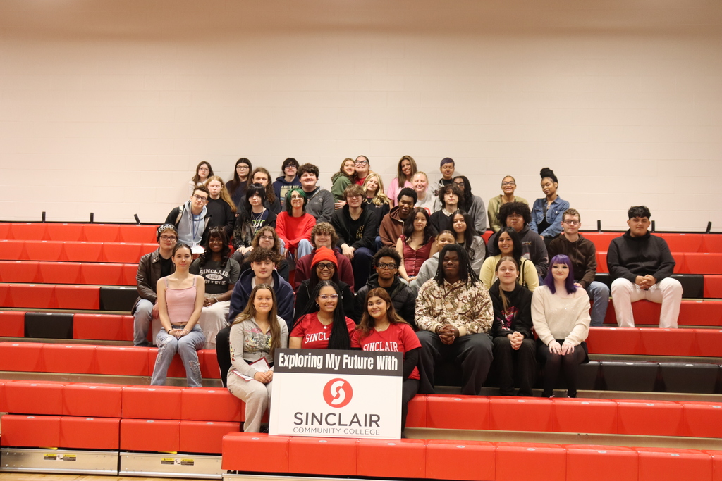 Group of students sits on red bleachers, some standing. Banner behind reads "Exploring My Future With Sinclair".