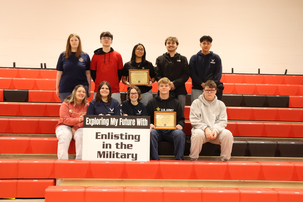 A group of students, some sitting and some standing, on red bleachers holding a sign that says "Exploring My Future, Enlisting in the Military"