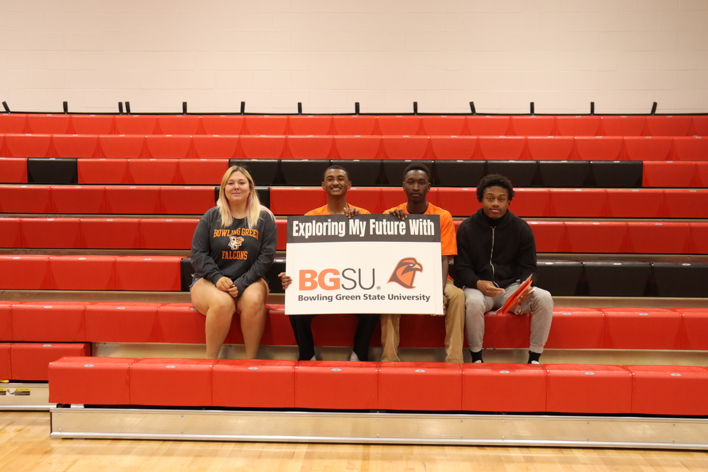 Four people are seated on a red bleacher. A sign displays "BGSU Exploring My Future With" and a bird logo.