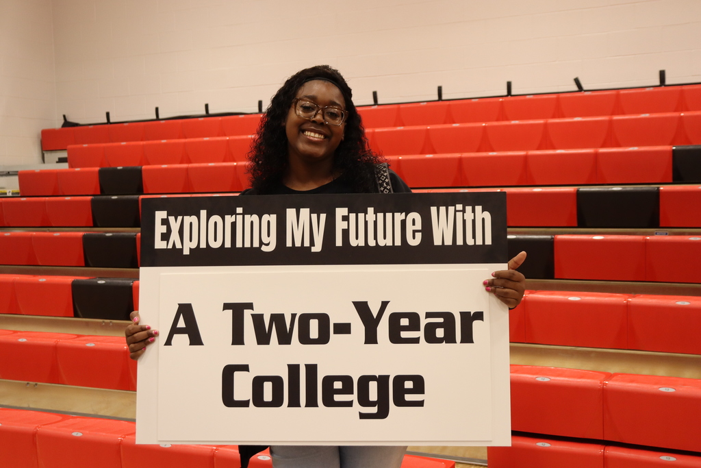 Person holding a sign that reads "Exploring My Future With A Two-Year College" in front of red stadium bleachers.