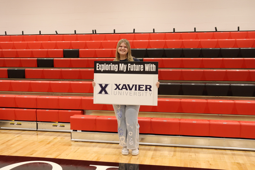 Woman holding sign "Exploring My Future With X XAVIER UNIVERSITY" in an indoor basketball court with red seating.