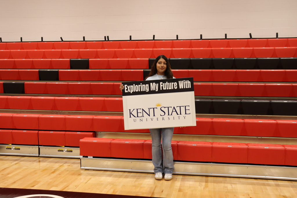 A woman holds a sign in front of a person, with "KENT STATE" on it. She stands in front of empty red bleachers.