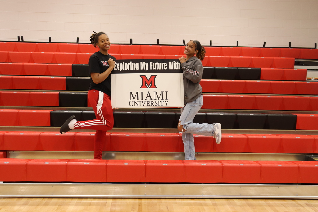 Two people stand on a red bleacher holding a sign reading "Exploring My Future with Miami University."