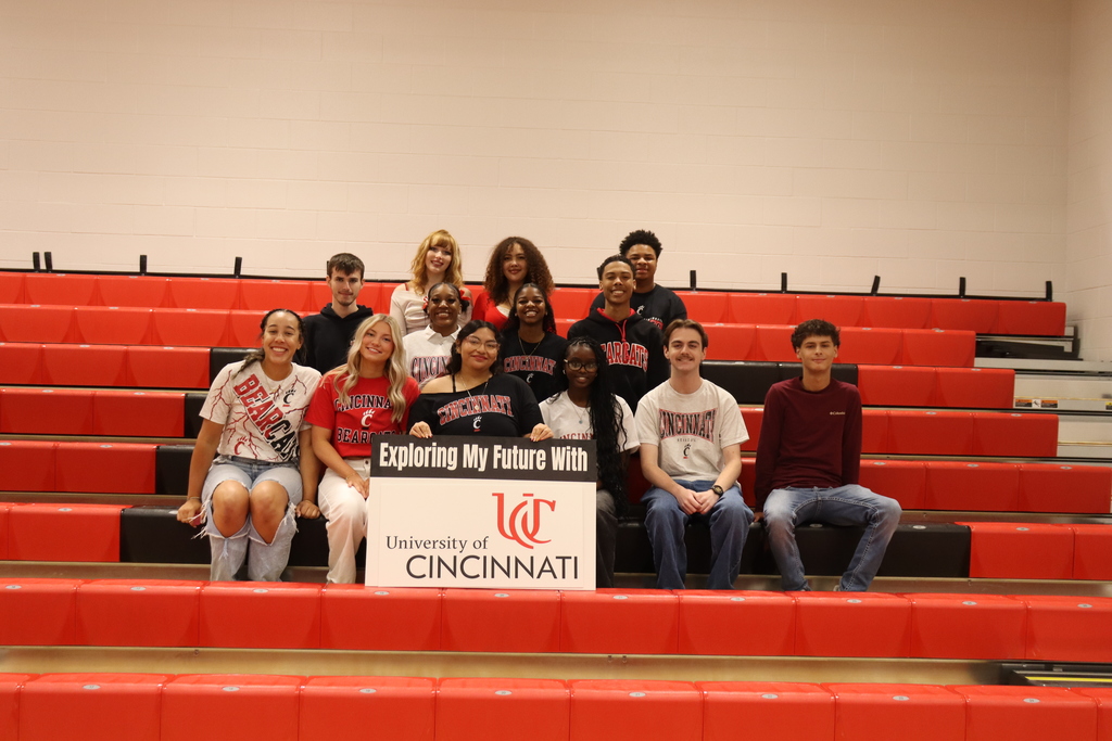 Group of people in a stadium, seated on red bleachers, holding a banner that reads "Exploring My Future with University of Cincinnati".