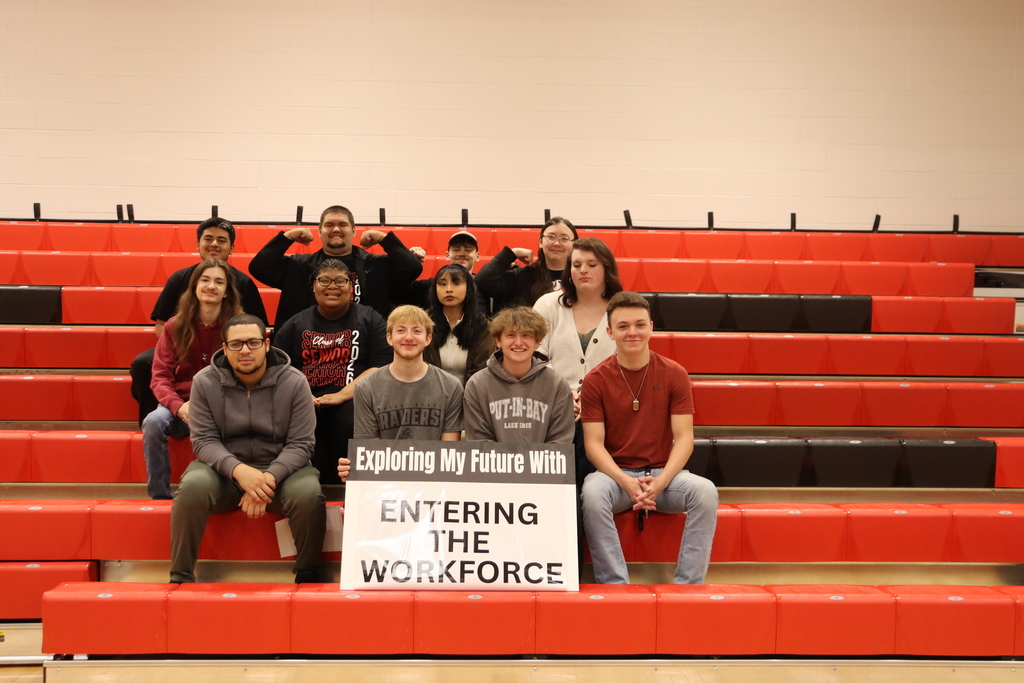 A group of individuals sits on red bleachers, holding a sign that reads "Entering the Workforce". They are seated in a row.