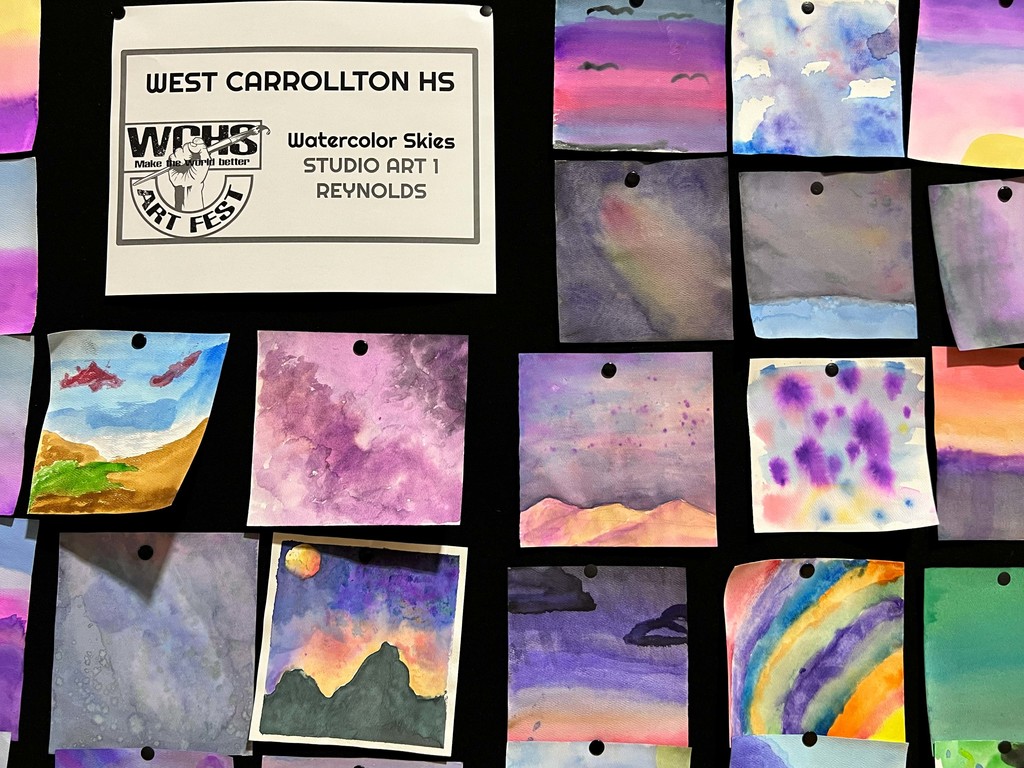 Multiple watercolor artworks pinned on a dark wall, including a sign that reads "Watercolor Skies Studio Art."