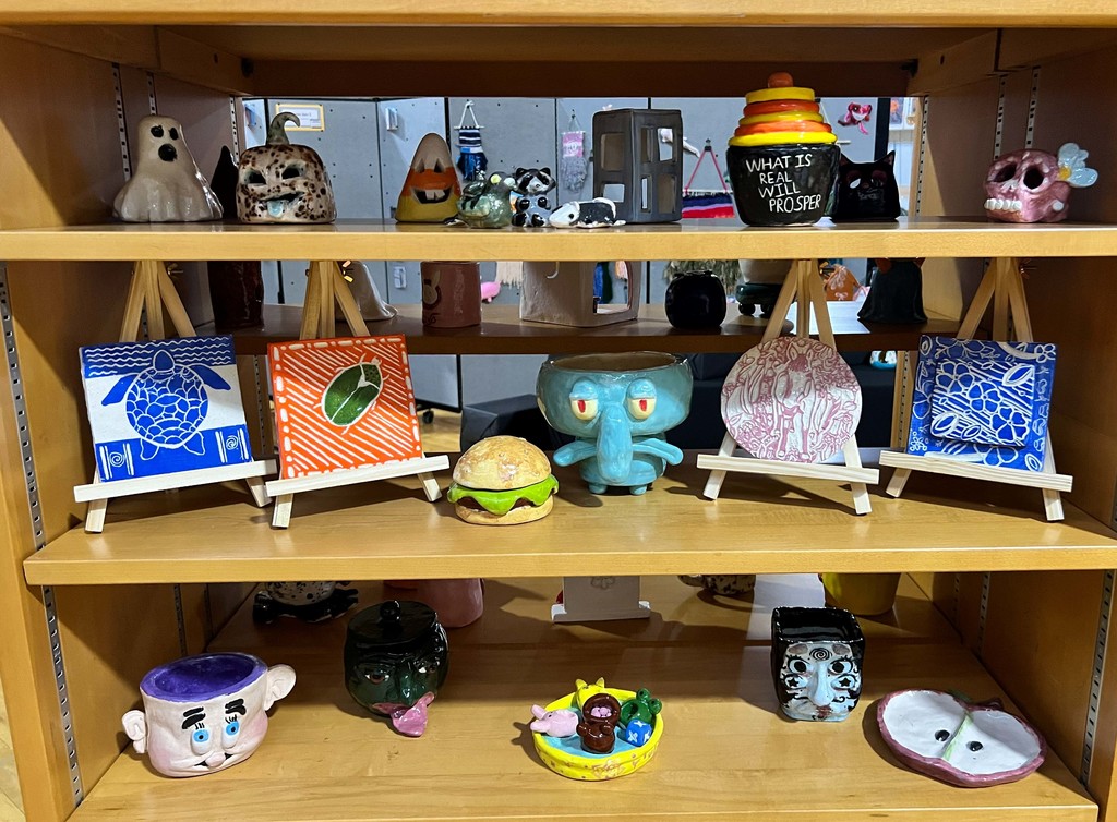 A wooden shelf displays figurines, miniature plates, and cups in various colors and designs. The figurines include a blue and white plate, a blue monster, a pink figurine, and a black cat.
