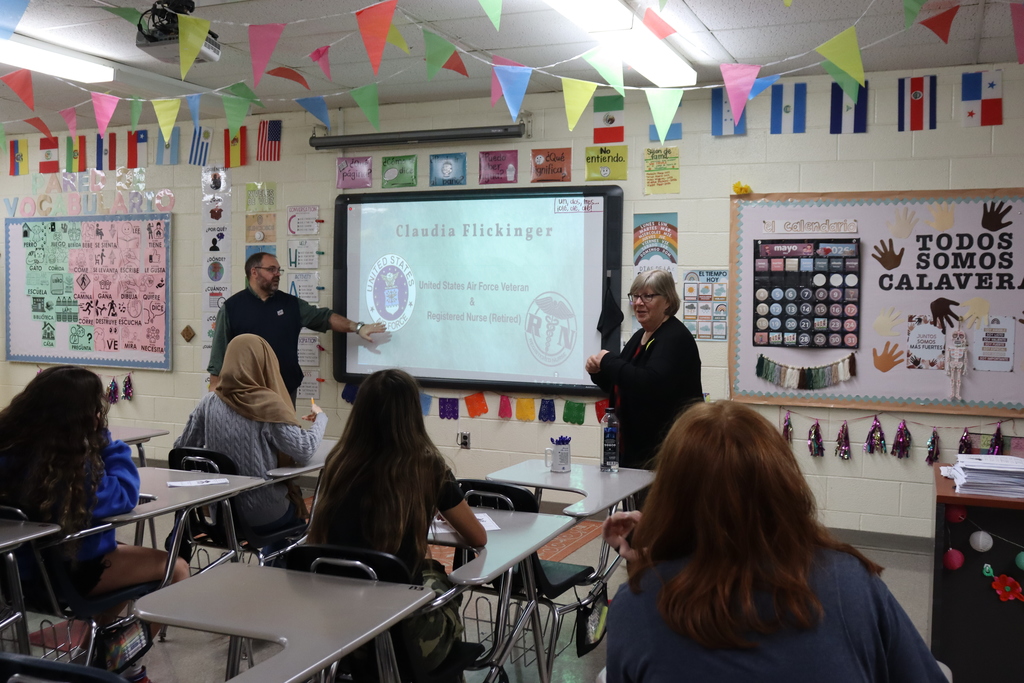 A classroom with students sitting at desks, a teacher presenting on a screen, and colorful decorations on the walls.