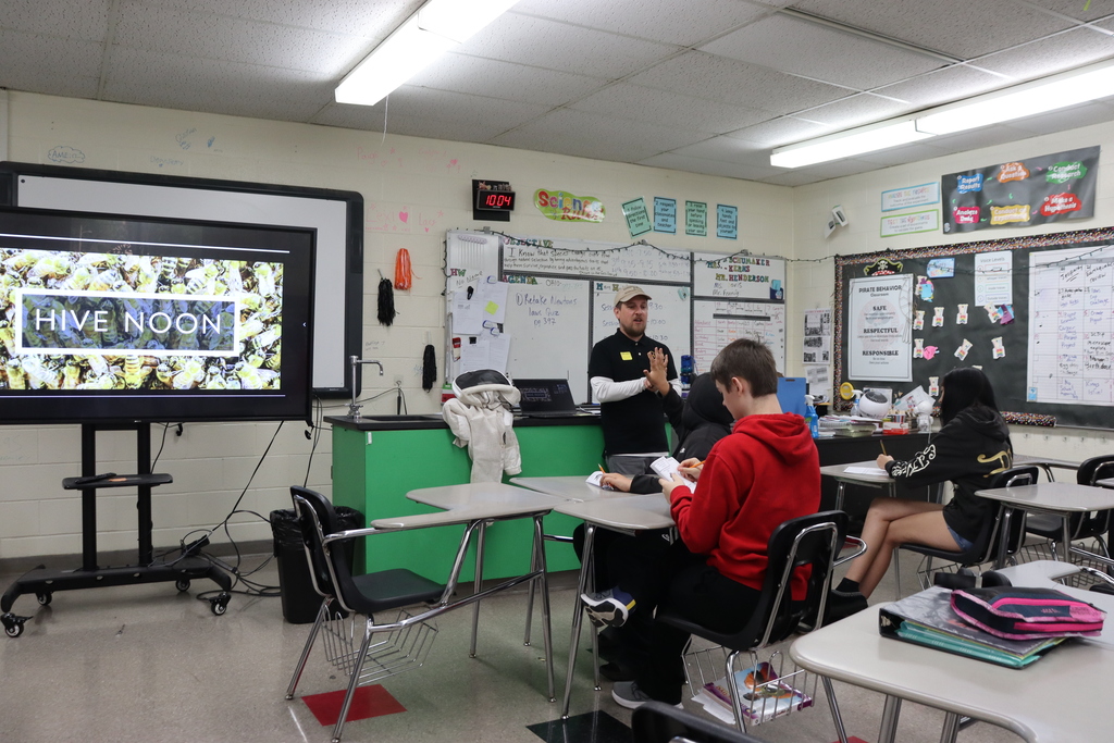 A classroom with a teacher standing by a screen. Students sit at desks. Whiteboards are on the wall.