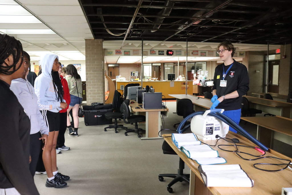 A group of individuals stands in a classroom; one person in a black shirt with a medical device. A table holds books, and computers sit on a nearby desk.