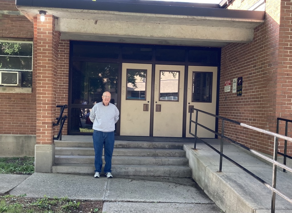 A person stands in front of a building with brick walls, two doors, and a staircase, wearing blue jeans and a gray sweater.