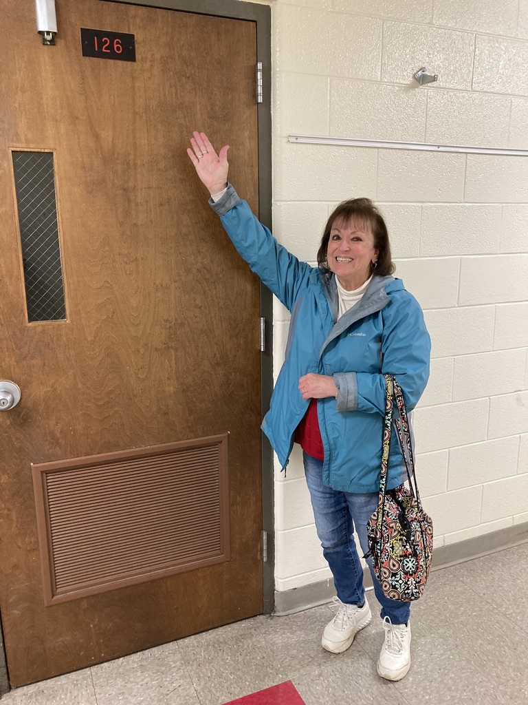 A woman with a colorful handbag waves while standing in front of a brown door.