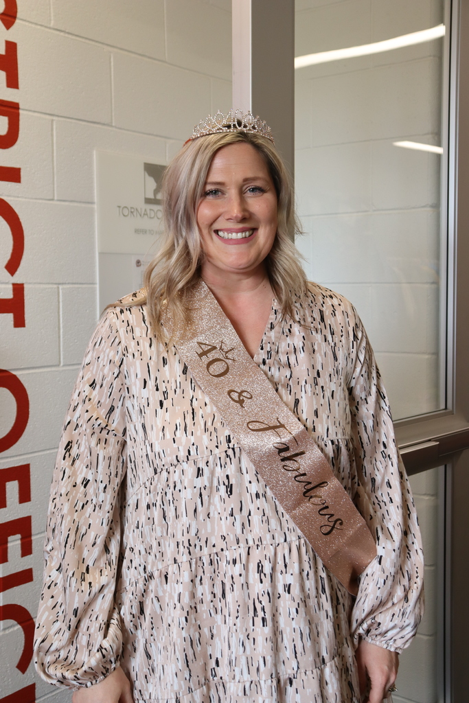 A woman wearing a dress and a sash reading "40 & Fabulous" stands smiling against a wall.