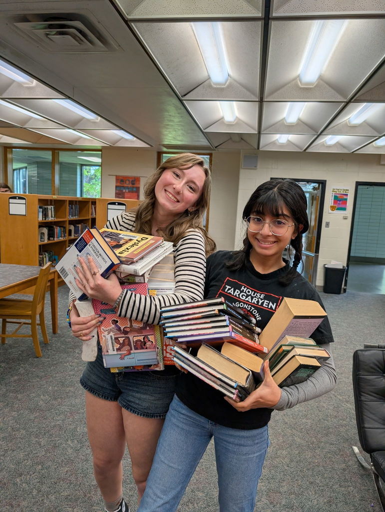 Two women stand in a library, holding large stacks of books. They smile and look at the camera.