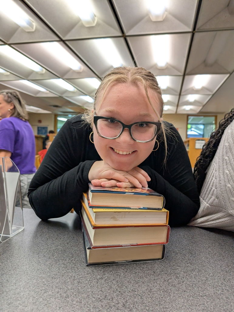 Woman sitting at a table in a library with a stack of books. She has glasses, a black shirt, and smiles.