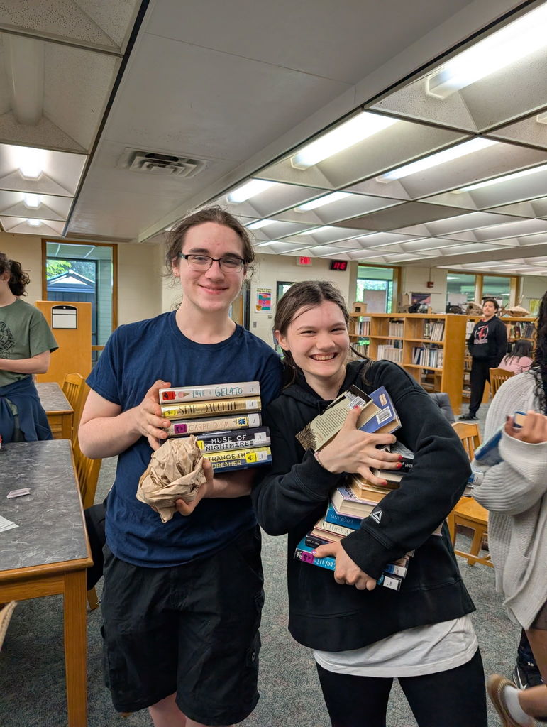 Two people in a library. One holds a stack of books, the other a bundle of books.