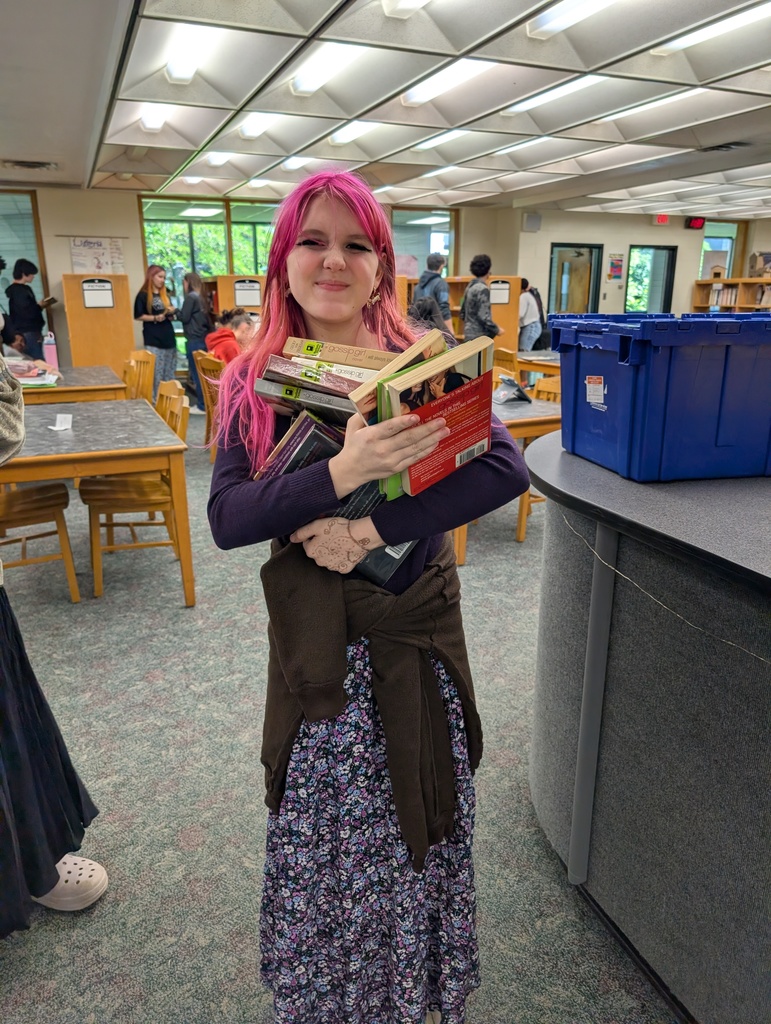 A woman with pink hair holds a stack of books in a library. Behind her, tables and chairs are visible, and a blue storage box rests on a counter.