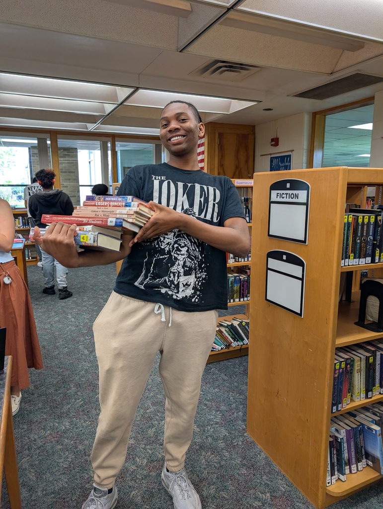 A young man holds a stack of books in a library, surrounded by bookshelves, with people in the background.