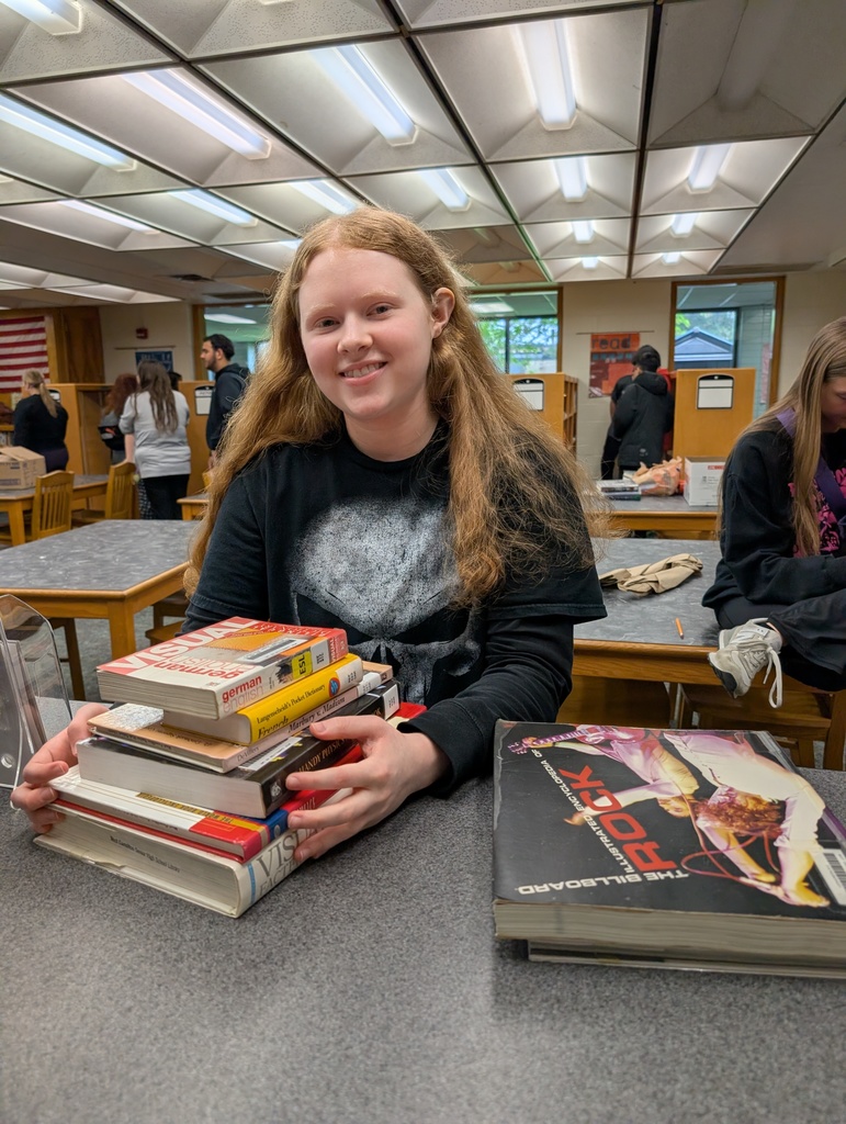 Person holding books, one with a rock design, in a library. Other students in the background.