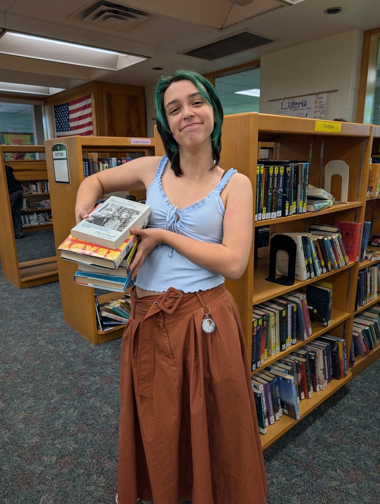 Woman holding a stack of books in a library. She wears a blue tank top and brown skirt.