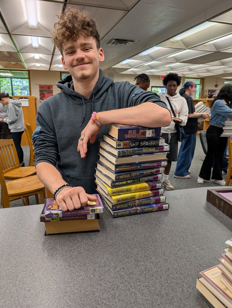 A man stands in a library, holding a stack of books, with other people and bookshelves in the background.