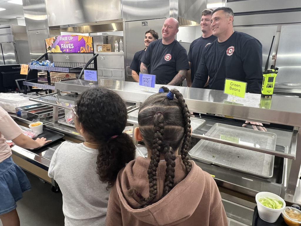 People in a cafeteria, two girls in front, trays of food on a counter, and four men in uniform behind.