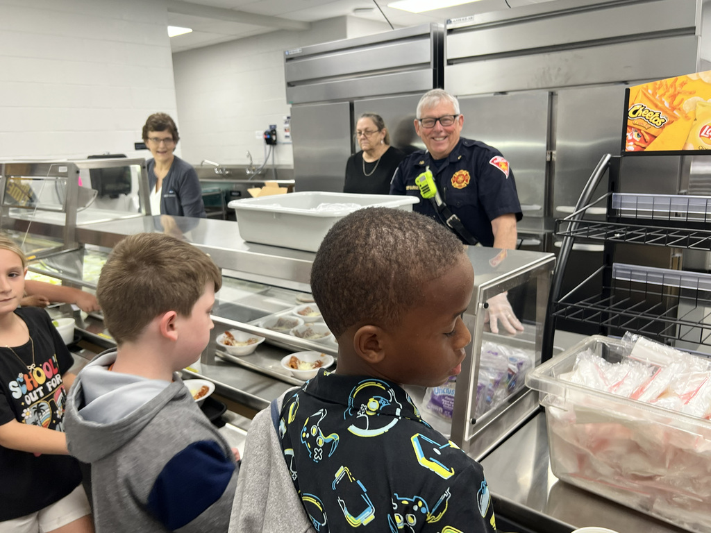 Several children stand near a serving counter, looking at the food. A firefighter and adults stand behind them.
