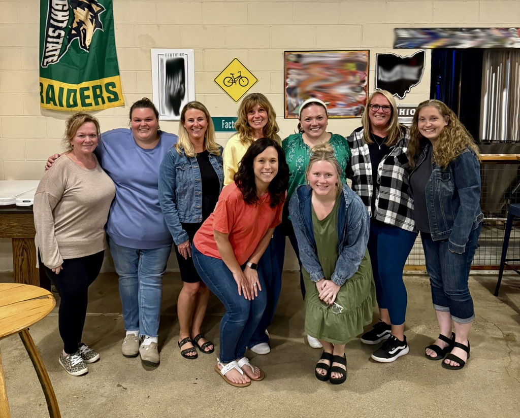 Nine women pose for a group photo in a room with a wall backdrop. Two women stand in front.