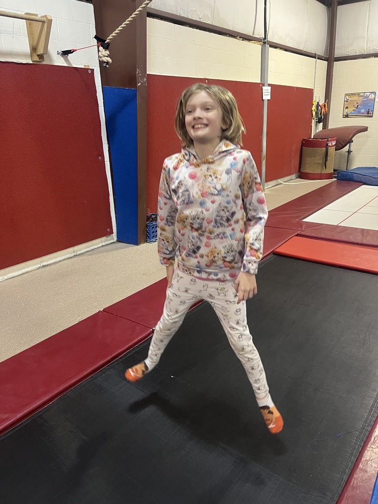 A young girl wearing a floral hoodie and pants, bouncing on a black trampoline in a gym.