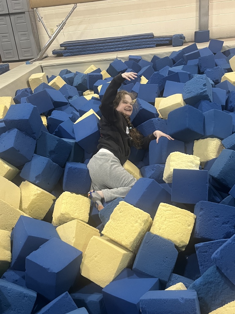A person in a black jacket and gray pants jumps in a foam pit filled with blue and yellow blocks.