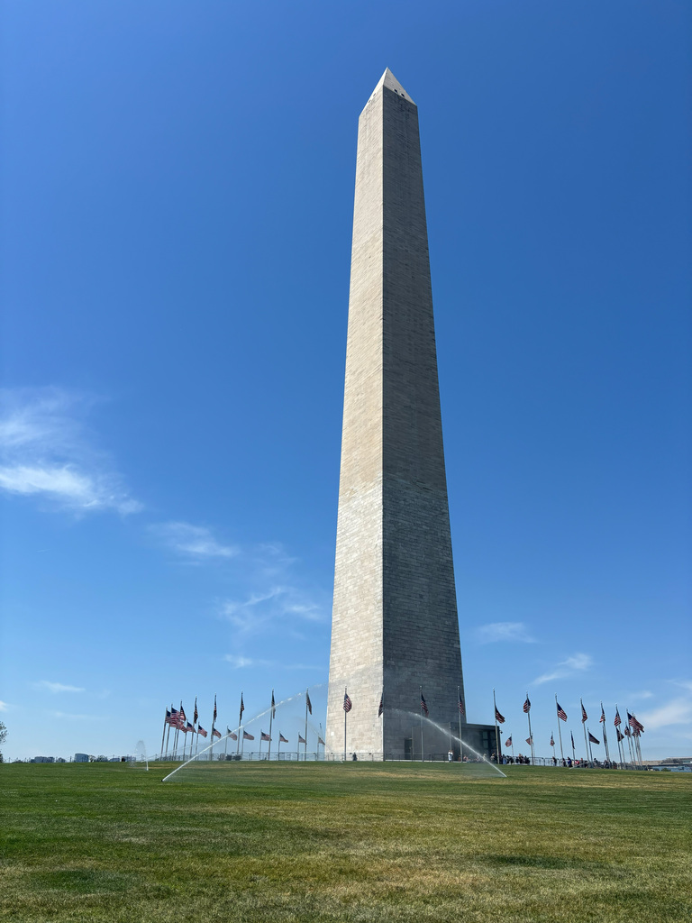 A tall, white obelisk stands tall against a blue sky. A green field with a sprinkler system in front.