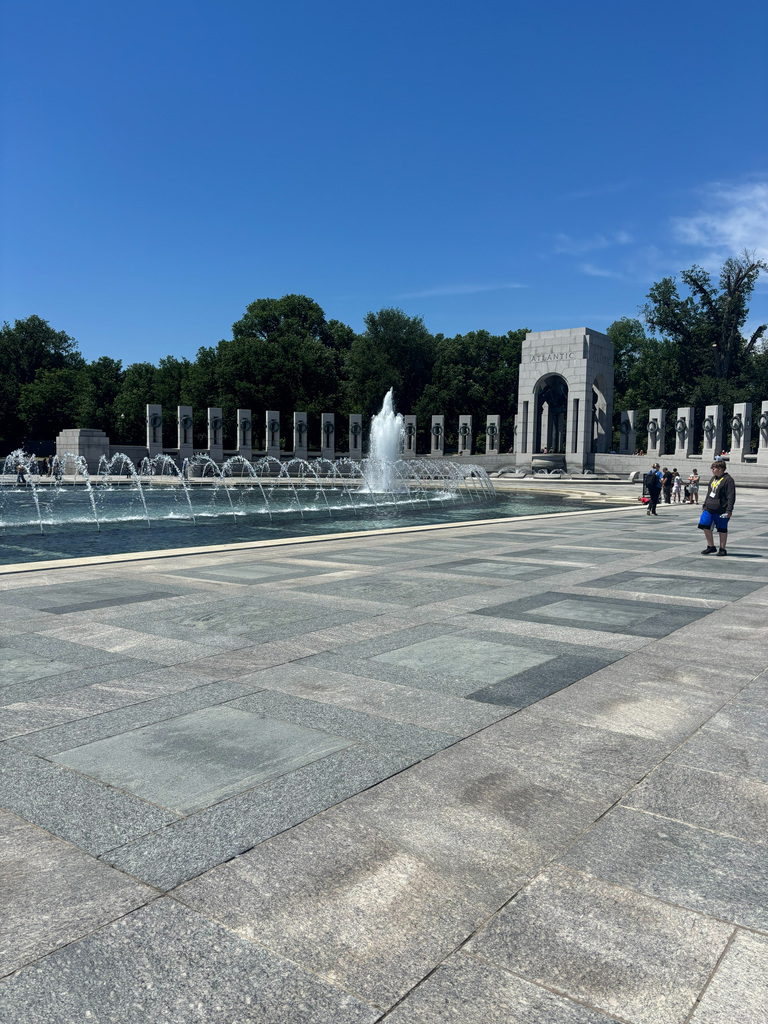 A memorial garden with a fountain in the center, surrounded by stone pillars. Visitors walk around. Trees are visible in the background.