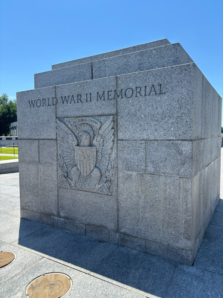 Gray stone wall, "World War II Memorial" engraved. An eagle, shield, and wings carved in the center. Bright sky above, shadows cast.
